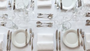 Ein vollständig edel gedeckter Tisch für mehrere Personen mit drei Gläser, Brotteller und -Messer, Besteck für jeden Gang und mittig eine Serviette.